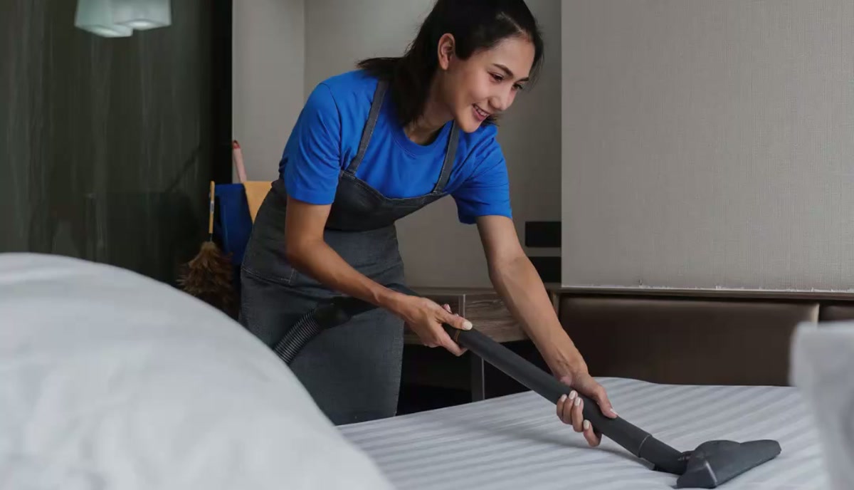 Professional cleaner vacuuming a bed in a hotel room